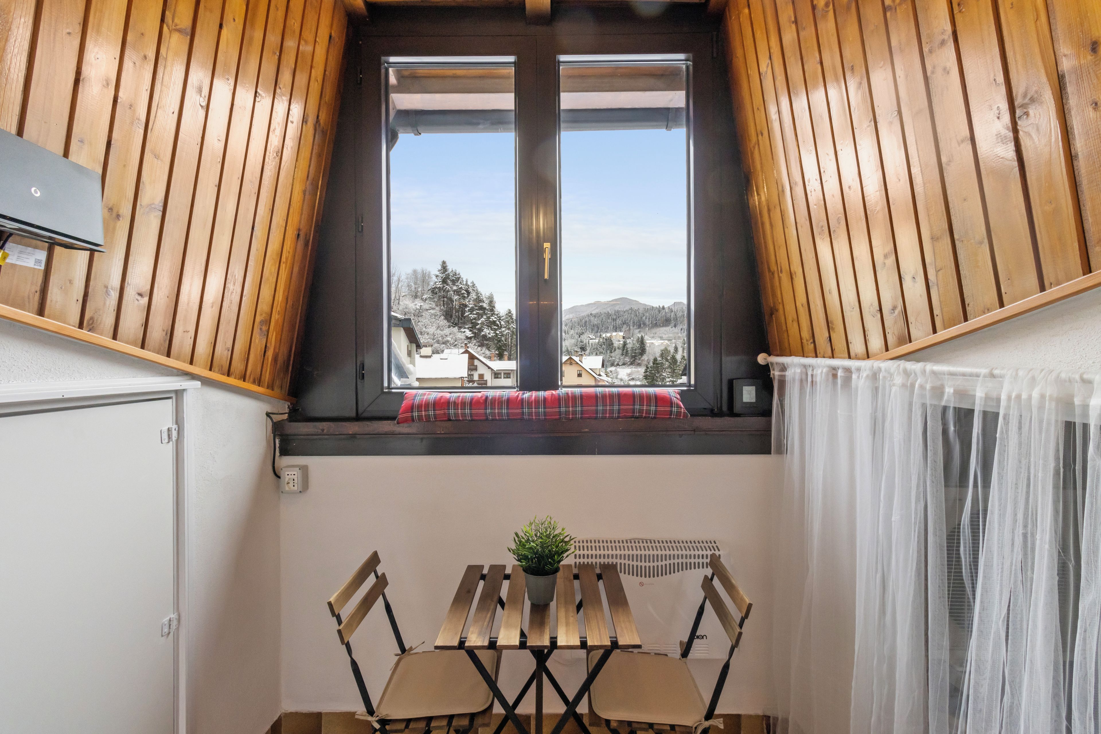 A room with a wooden sloped ceiling, a window with a view of snow-covered trees and buildings, a small table with chairs, and a white curtain.