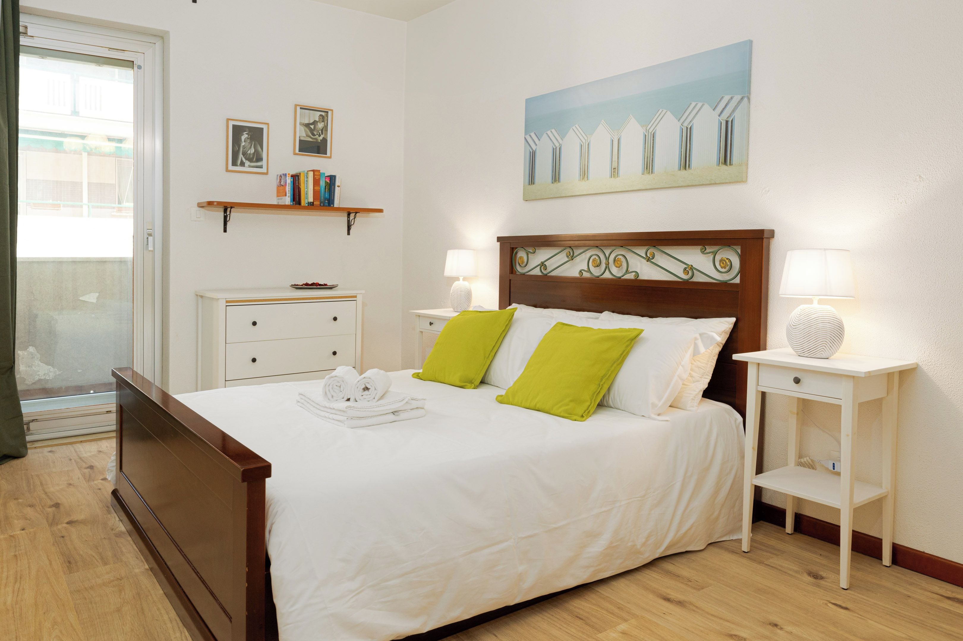 Una cama bien hecha con almohadas, una mesita de noche con lámpara, un mueble auxiliar y un estante en la pared con libros.