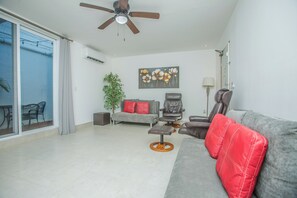 A modern living room with a grey sofa, a brown leather chair, a small table, a potted plant, and a ceiling fan. - Jaguar Condos Cozumel The Private Group Villa / Casa 1-24 w/2 pools Downtown (Cozumel)