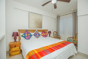 Ein Hotelzimmer mit einem Bett, einem Deckenventilator, einem Nachttisch aus Holz, einer Holzkommode und einem Fenster mit Vorhängen.