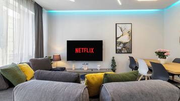 A modern living room with a flat-screen TV displaying 'NETFLIX,' a dining area with a wooden table and chairs, and a decorative plant.