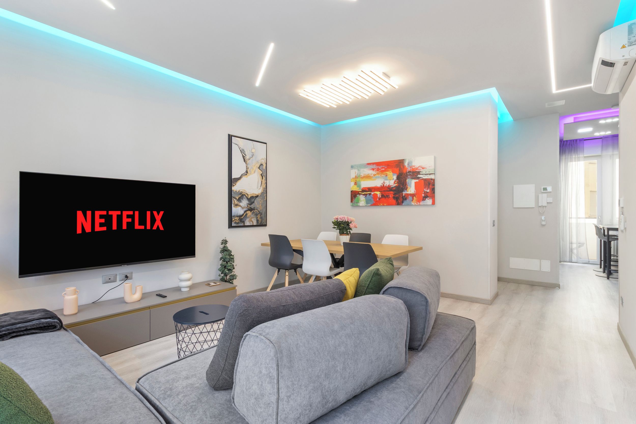 A modern living room with a flat-screen TV displaying 'NETFLIX,' a sofa, a coffee table, and a dining area with chairs and a table.