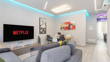 A modern living room with a flat-screen TV displaying 'NETFLIX,' a sofa, a coffee table, and a dining area with chairs and a table.