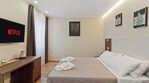 Une chambre d’hôtel avec un lit, un bureau doté d’une bouilloire, un téléviseur affichant le logo « NETFLIX », et deux œuvres d’art encadrées accrochées au mur.