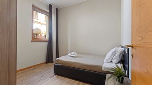 Una stanza piccola con un letto singolo, pavimento in legno, una finestra con tende, una pianta sul comodino e una porta con maniglia in ottone.