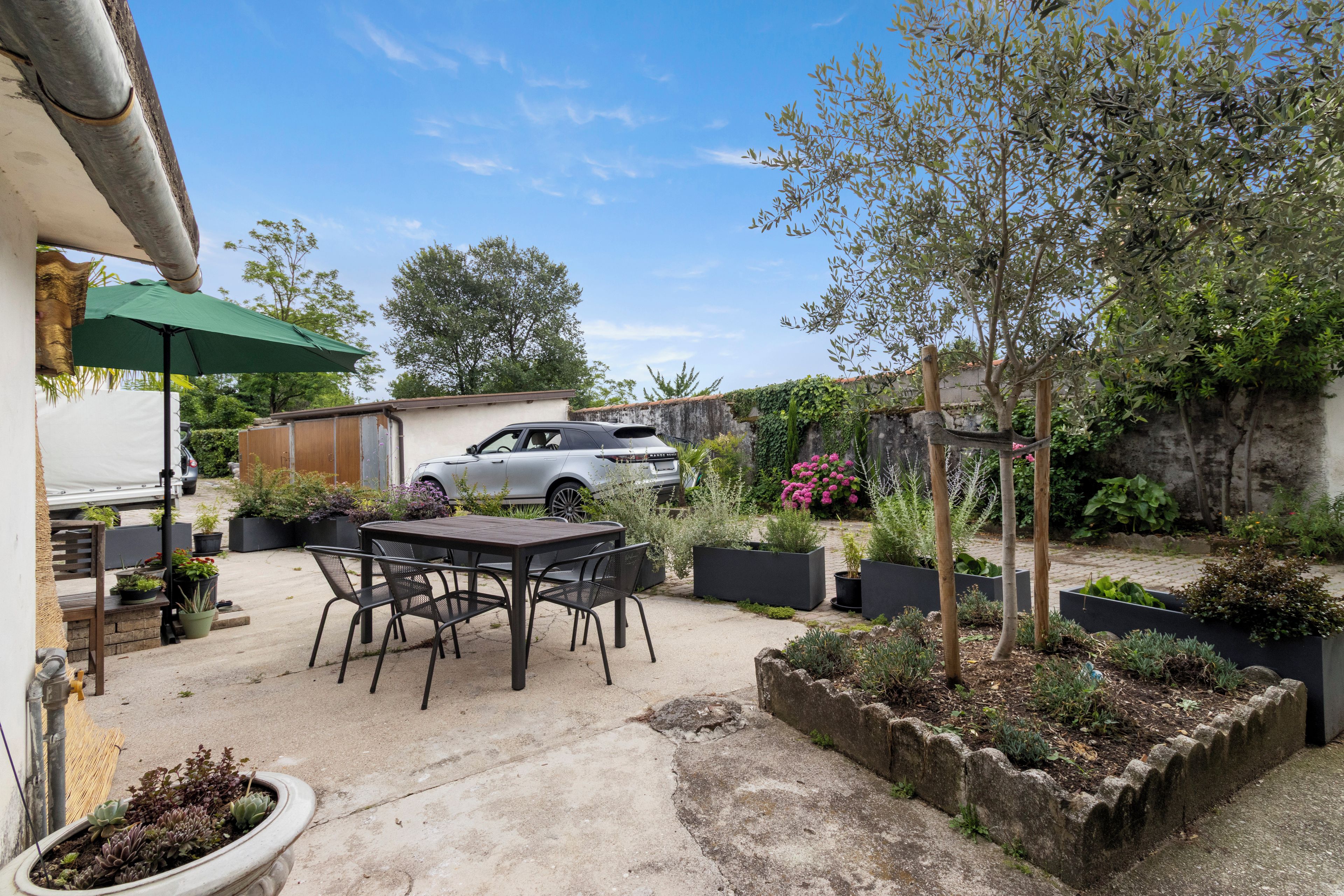 A patio area with a table and chairs, surrounded by planters and a car parked in the background.