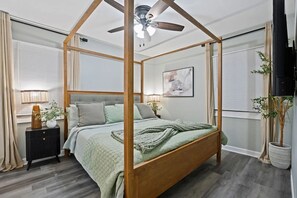 Un llit amb cortines, un ventilador al sostre, una tauleta de nit amb una làmpada i una planta en test.