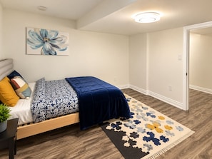 Un dormitorio con cama, mesita de noche, lámpara, una obra de arte en la pared y una puerta.