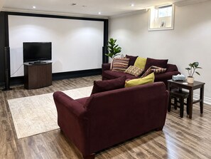 A living room with a dark-colored sofa, a flat-screen TV on a stand, a small table with a plant, and a large window. - Private Suite | Sauna, Cinema, Gym & Pool Table (King)