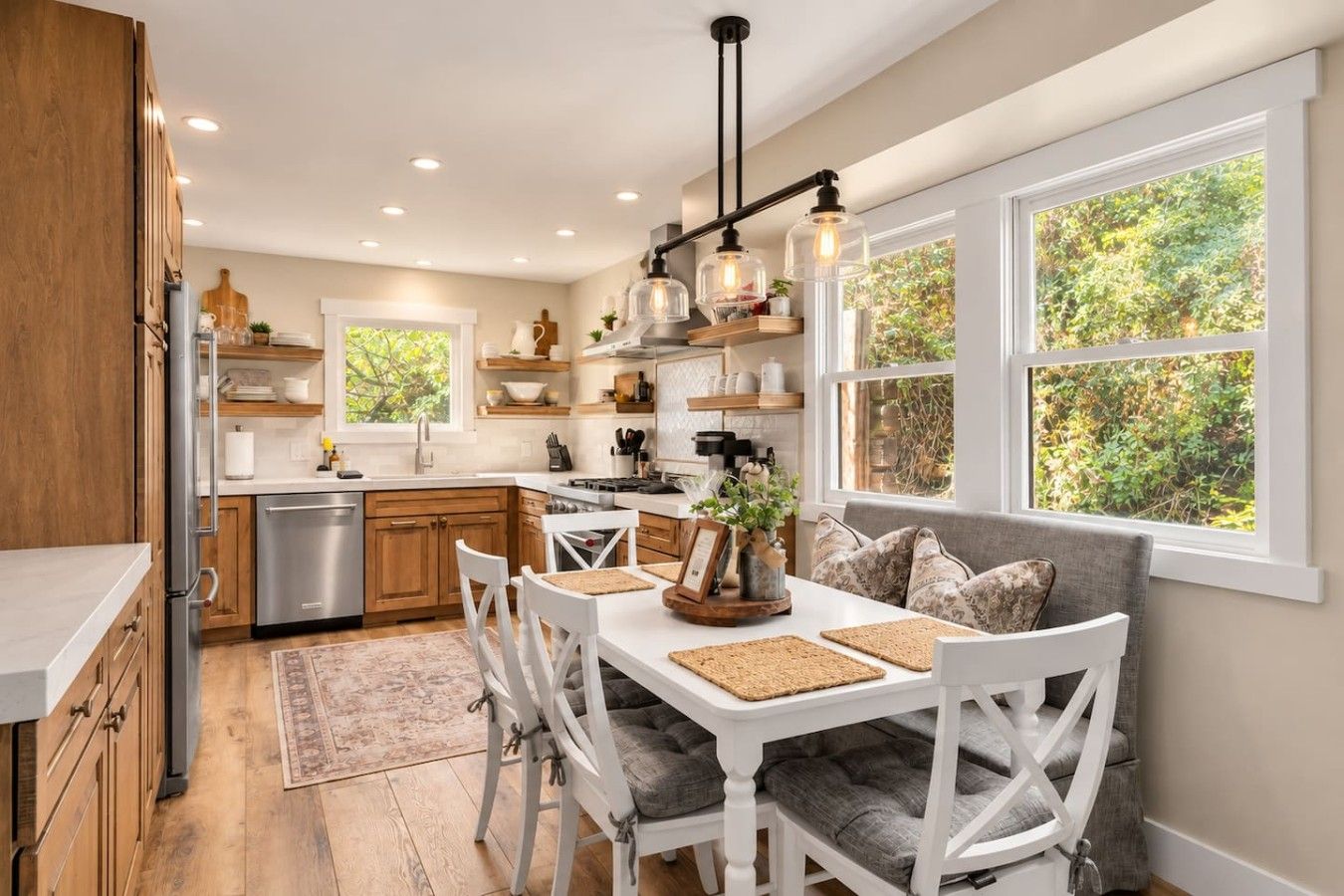 A modern kitchen with white cabinetry, a dining area with a white table and grey upholstered chairs, and large windows with a view of greenery.