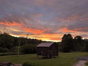 Une structure rustique en bois, située dans un champ verdoyant, avec un ciel au coucher du soleil.