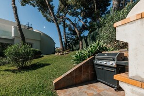 Eine Terrasse mit Grill, ein weißes Gebäude und eine begrünte Fläche mit Bäumen.