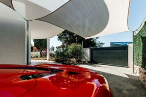 Ein roter Sportwagen steht unter einer großen Markise, im Hintergrund eine Garagentür und viel Grün.