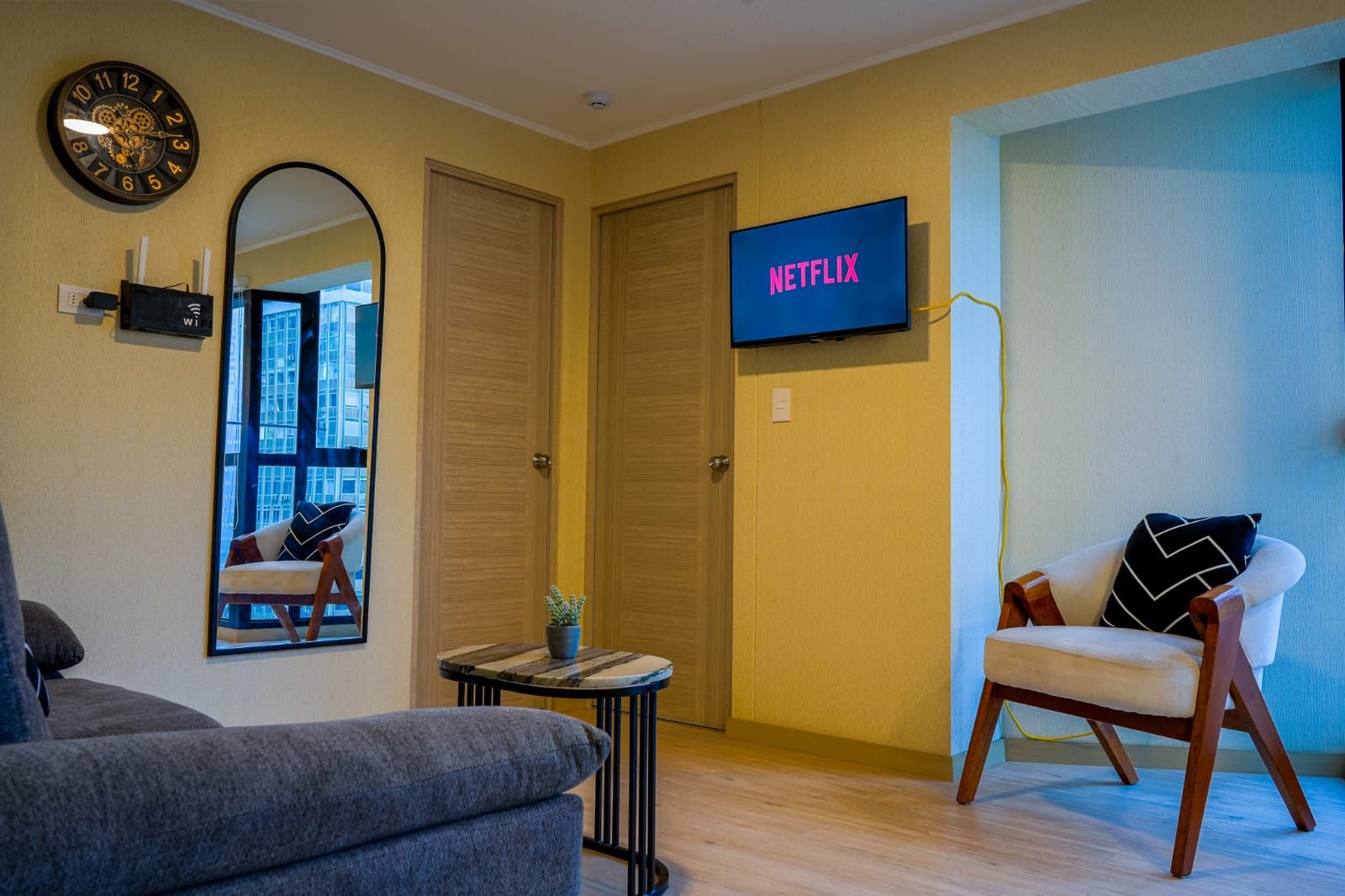 A modern living room with a sofa, a chair, a clock, a mirror, a small table, and a TV displaying 'NETFLIX'.