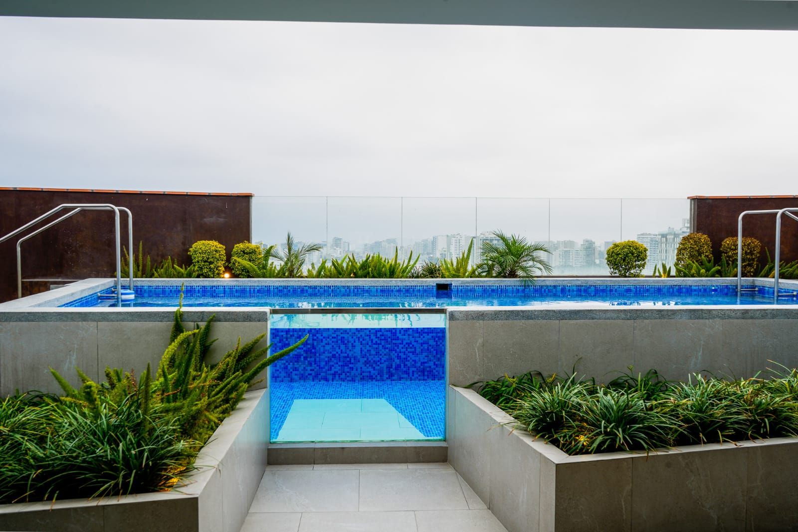 A rooftop swimming pool with a city view, surrounded by greenery and concrete structures.