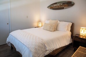 A neatly made bed with a white comforter and pillows, a bedside lamp, and a wall sign that reads 'The BEACH is my HAPPY PLACE.' - Tide · Beach days, family nights (Cocoa Beach)