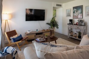 Ein Wohnzimmer mit Flachbildfernseher, einer Couch, einem Couchtisch, einer Stehlampe, einem Bücherregal und einem Topf mit Pflanze.