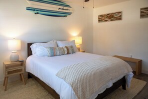 A bedroom with a bed, two bedside lamps, a nightstand, a decorative surfboard, and a wall art piece. - Sunset · Beach days together | 2BR, sleeps 6 (Cocoa Beach)