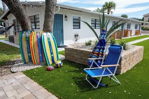 A house with a front yard, a blue door, and a small patio area with a blue lawn chair. - Coral House · Whole House · 3 Suites · Sleeps 12 (Cocoa Beach)