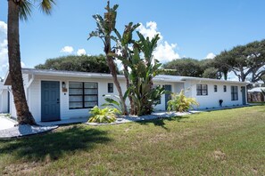 A single-story building with a white exterior, a blue door, and a small front yard with a palm tree and other tropical plants. - Coral House · Whole House · 3 Suites · Sleeps 12 (Cocoa Beach)