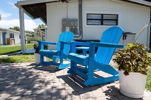 Eine Terrasse mit zwei blauen Adirondack-Stühlen, einem Blumentopf und einem weißen Gebäude mit einem Fenster und Schaltkästen.