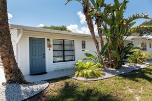 Einbungalow mit dunkelblauer Haustür, einer kleinen Veranda und einem gepflegten Garten mit Palmen und einem Kiesweg.