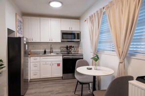 A compact kitchen with white cabinets, a black refrigerator, a microwave, and an oven. There's a dining table with two chairs and a small plant on it. - Garnet Beaches · 2 Houses · 6 Suites · Sleeps 24 (Cocoa Beach)