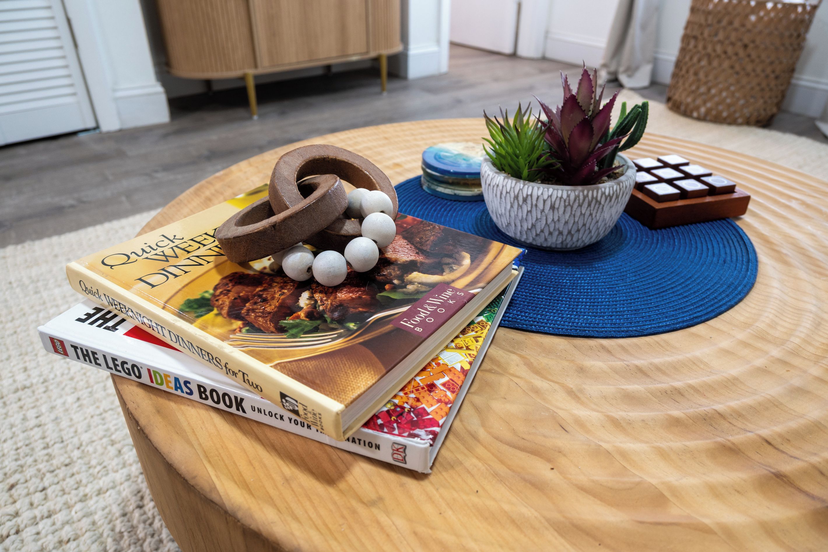 A stack of cookbooks on a wooden table with a blue placemat, a potted plant, and a wooden puzzle.