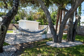 Eine Hängematte zwischen Bäumen, ein Geräteschuppen und eine Terrasse mit Stühlen und Tisch.
