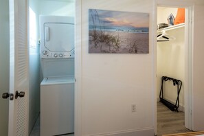 A laundry room with a washing machine and dryer, a painting of a beach scene, and a shoe rack in the adjacent room. - Sand · Pack light, stay awhile | 2BR + laundry (Cocoa Beach)
