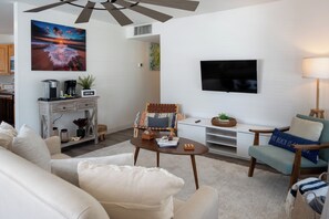 A modern living room with a flat-screen TV, a coffee machine, and a comfortable seating area with a sofa and armchairs. - Sand · Pack light, stay awhile | 2BR + laundry (Cocoa Beach)