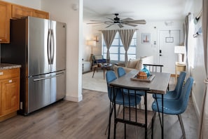 A modern kitchen with a stainless steel refrigerator, wooden cabinets, and a dining area with blue chairs and a wooden table. - Sand · Pack light, stay awhile | 2BR + laundry (Cocoa Beach)