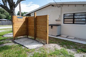A wooden shower enclosure with a sign, a small shed, and a house with a window and electrical box. - Sand · Pack light, stay awhile | 2BR + laundry (Cocoa Beach)