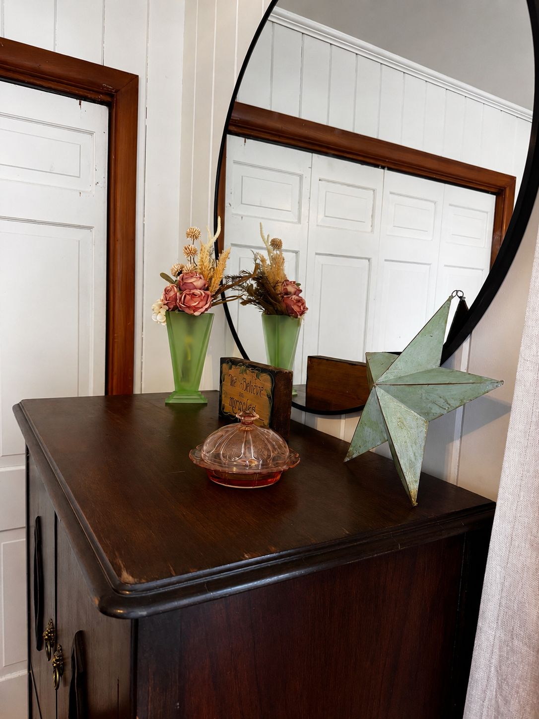 A wooden dresser with a mirror, two green vases with flowers, a small sign, and a geometric paper decoration.