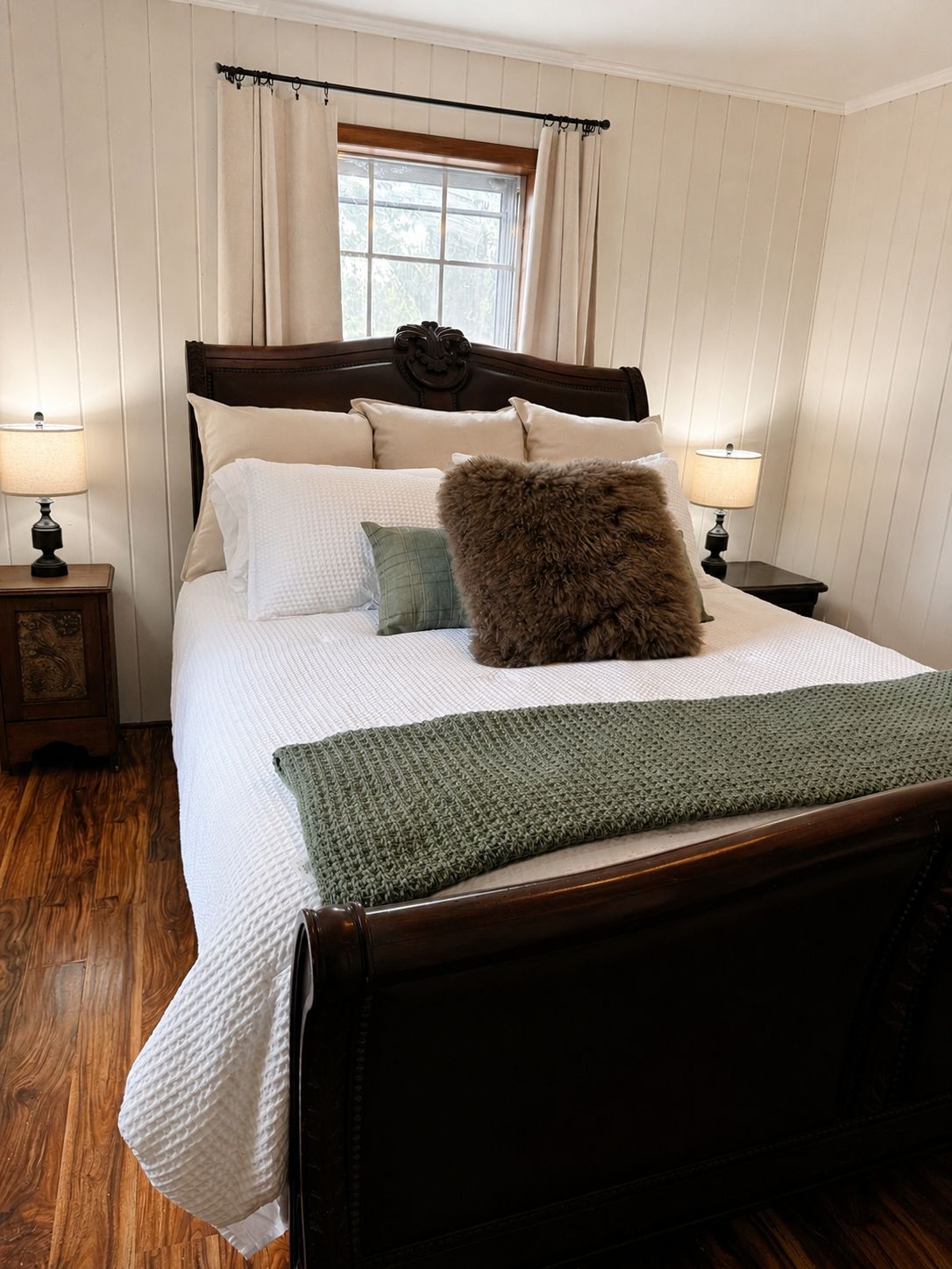 Una cama bien hecha con cabecera de madera, dos mesitas de noche con lámparas y una ventana con cortinas.