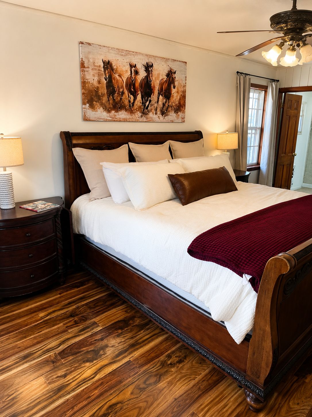 Una cama bien tendida con cabecera, piso de madera, pintura de caballos y un ventilador de techo.