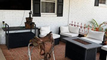 Un patio al aire libre cubierto con piso de ladrillo, paredes de ladrillo blanco, un banco de metal negro, un sofá blanco y una mesa de metal con hogar.