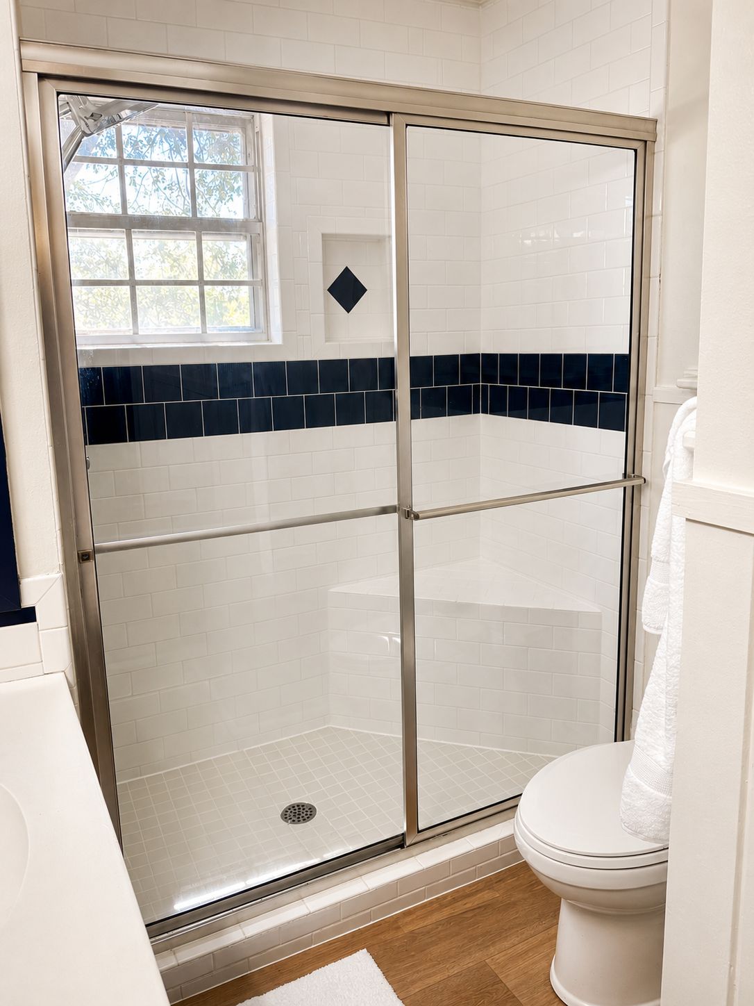 A bathroom with a glass-enclosed shower, white tiles, and a wooden floor.