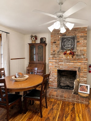 A dining area with a brick fireplace, wooden table, chairs, and a ceiling fan.