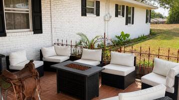Un patio cubierto con mobiliario exterior, una fogata y un edificio de ladrillo blanco al fondo.