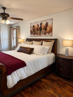 A bedroom with a large bed, a ceiling fan, a painting of horses, a nightstand, and a lamp.