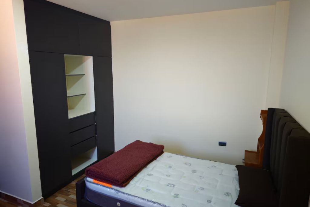 A small, compact bedroom with a single bed, a built-in wardrobe with multiple shelves, and a plain wall.