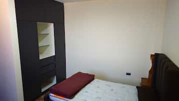 A small, compact bedroom with a single bed, a built-in wardrobe with multiple shelves, and a plain wall.