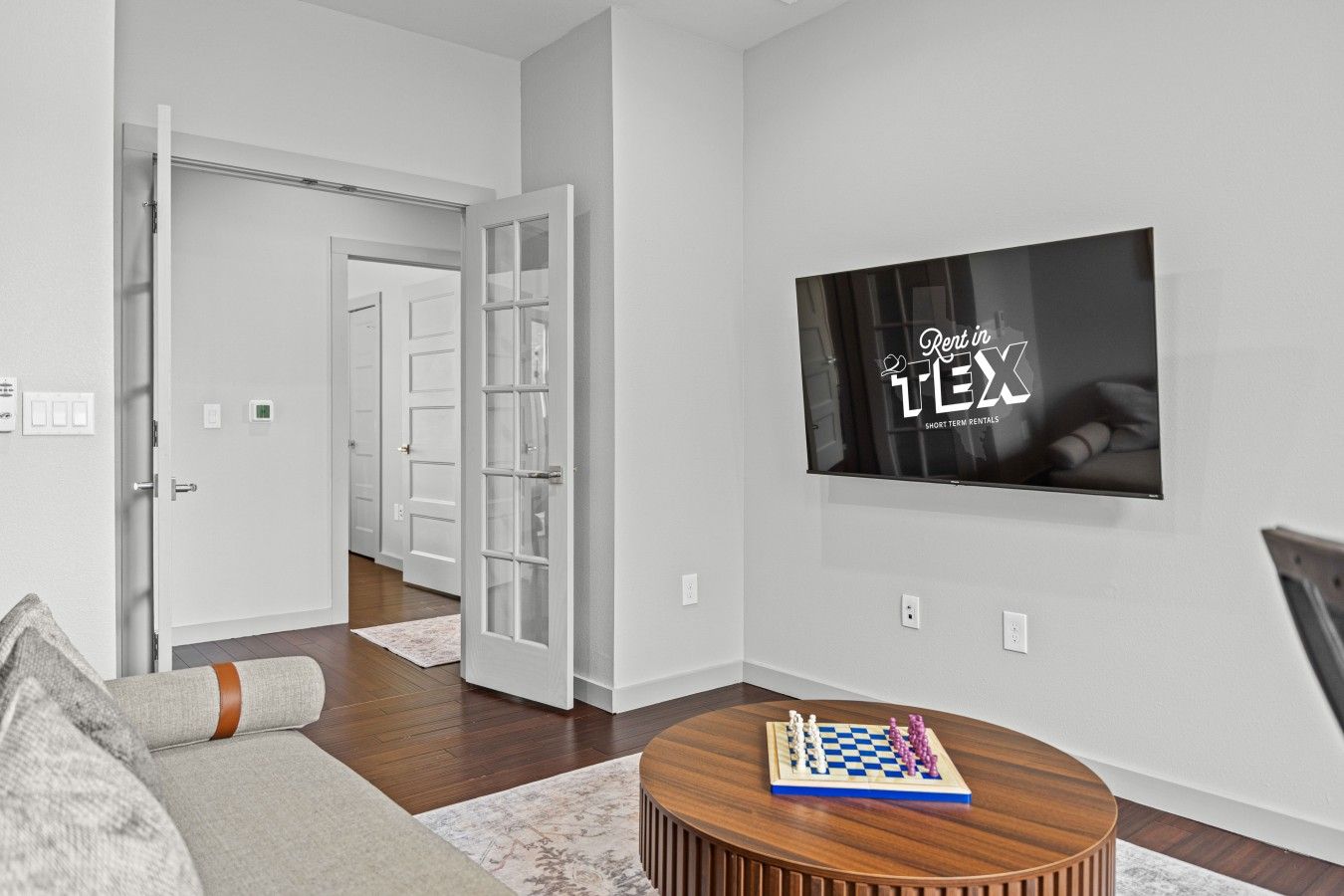 A modern living room with a flat-screen TV mounted on the wall, a wooden coffee table with a chess set, and a hallway leading to a door.