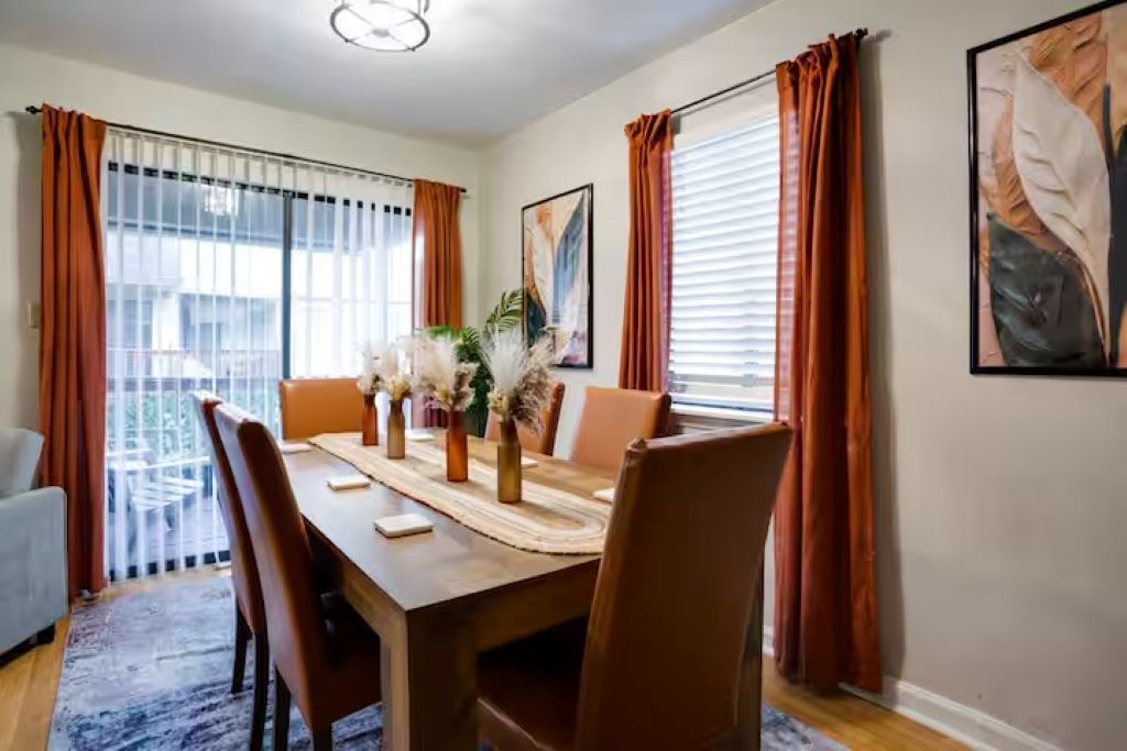 Uma sala de jantar com uma mesa de madeira, cadeiras e um vaso com plantas secas.