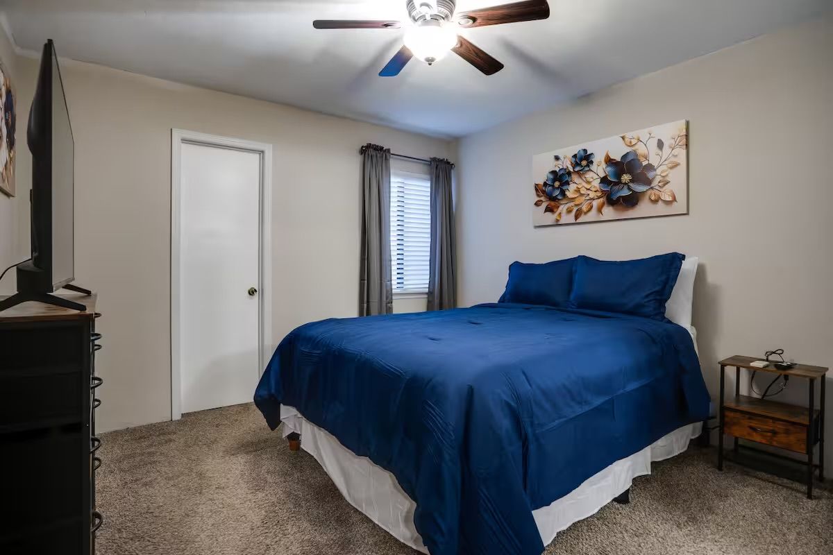 A bedroom with a bed, a ceiling fan, a door, a window with curtains, and a wall-mounted artwork.