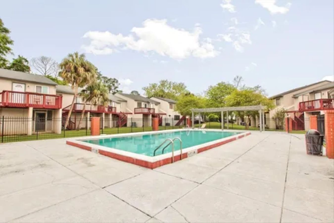 Piscina com borda vermelha, cercada por área pavimentada com um espaço de estar coberto e um edifício com varanda vermelha.