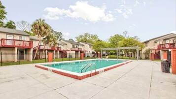 Piscina com borda vermelha, cercada por área pavimentada com um espaço de estar coberto e um edifício com varanda vermelha.