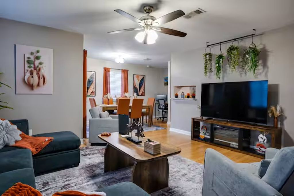 Sala de estar com sofá, mesa de centro e televisão. Há uma área de jantar com cadeiras e uma mesa ao fundo.