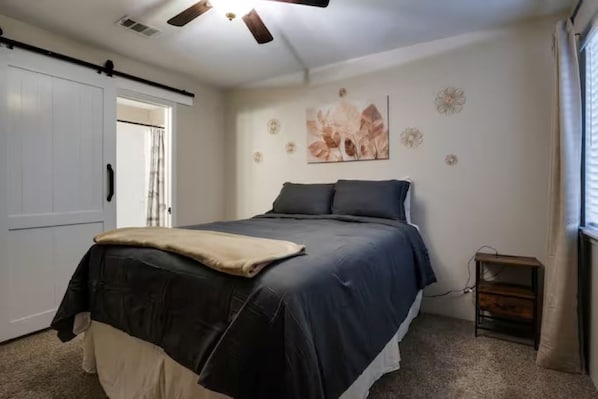A bedroom with a bed, a ceiling fan, a door, and a small table.